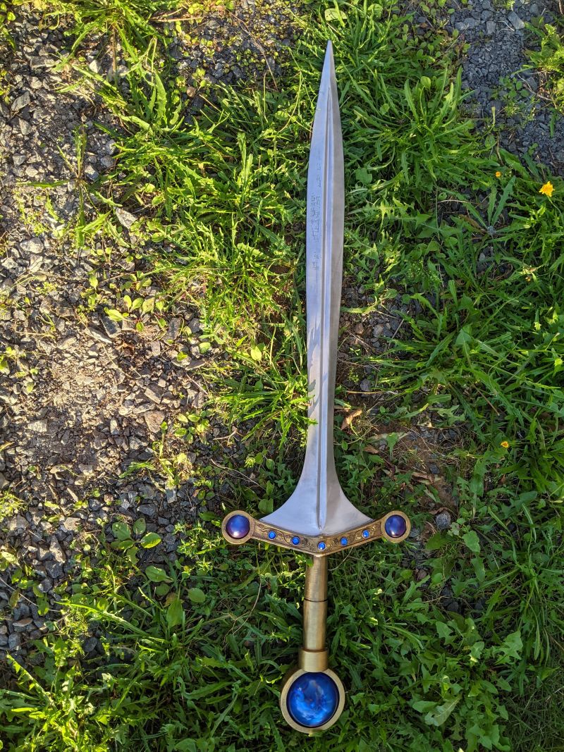 A long flared sword with a massive blue sapphire pommel, two large blue saphires on each end of aa golden guard inlaid with grape vines, The blade has sanksrit inscribed on it.